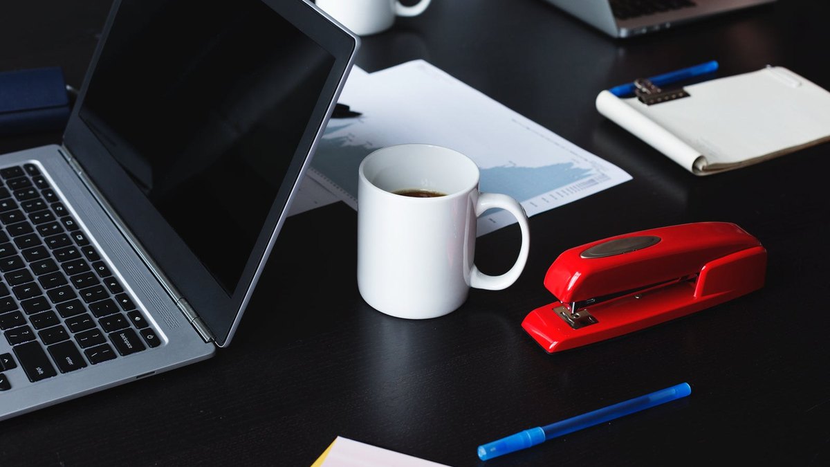 A laptop, coffee mug, red stapler and small notebook on a dark meeting table.