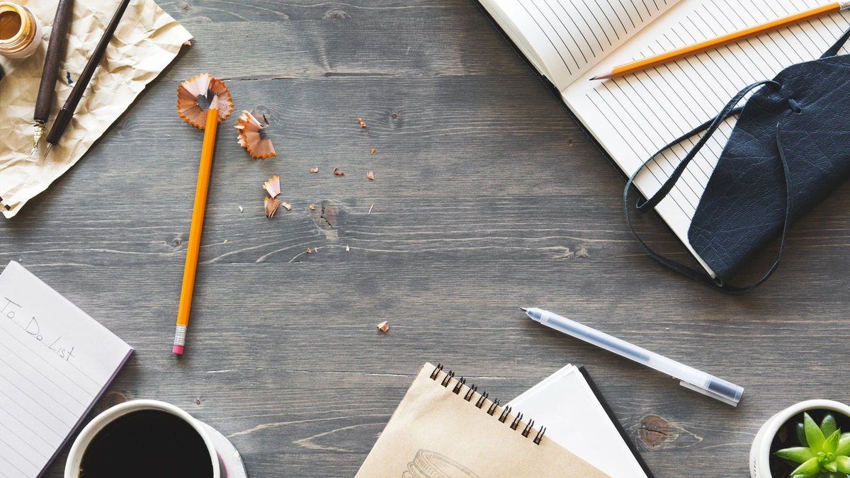 Pencils, a pen, notebooks and pencil shavings on a grey wooden desk.
