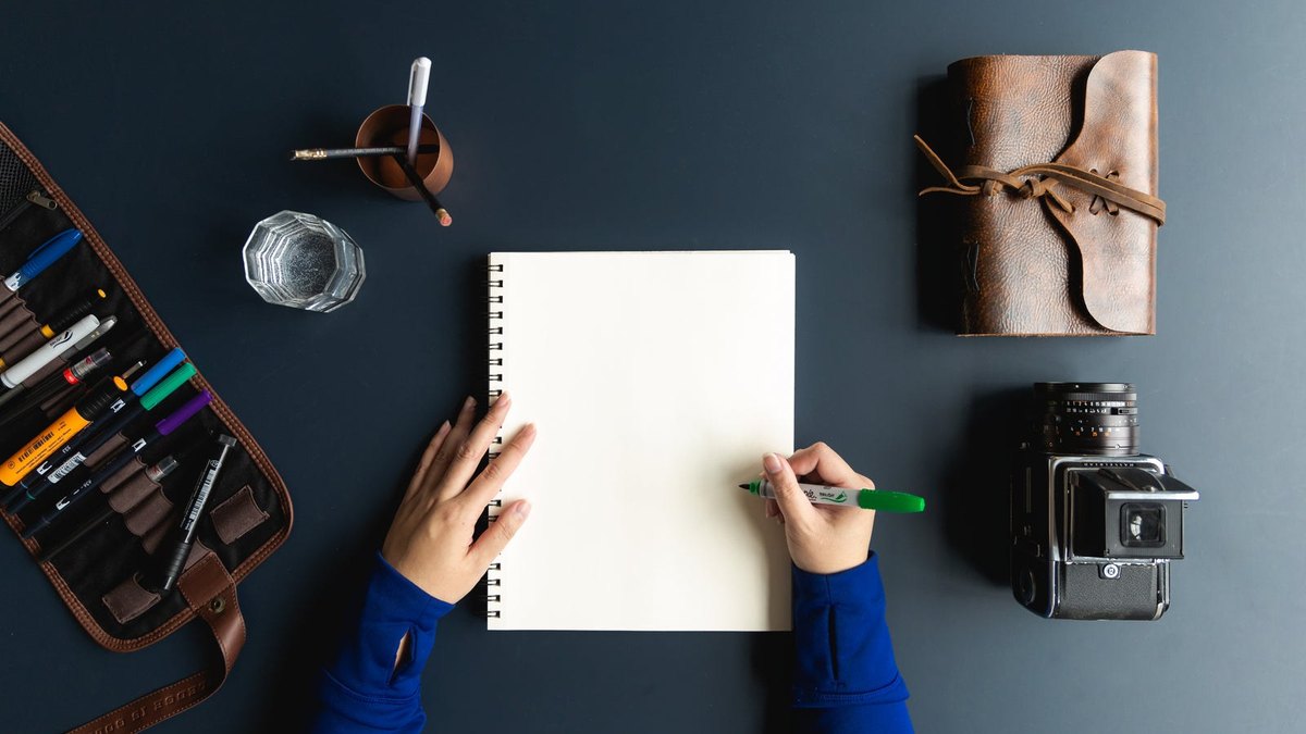 Hands holding a blank notebook and green pen on a dark desk with writing materials.