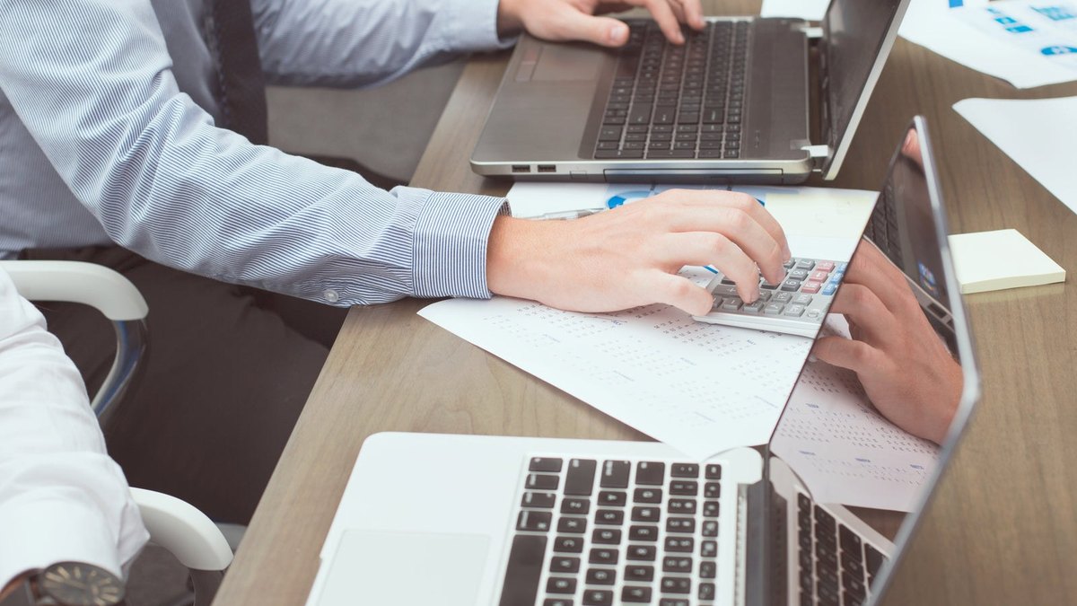 A hand using a calculator beside paperwork and laptops on a meeting table.