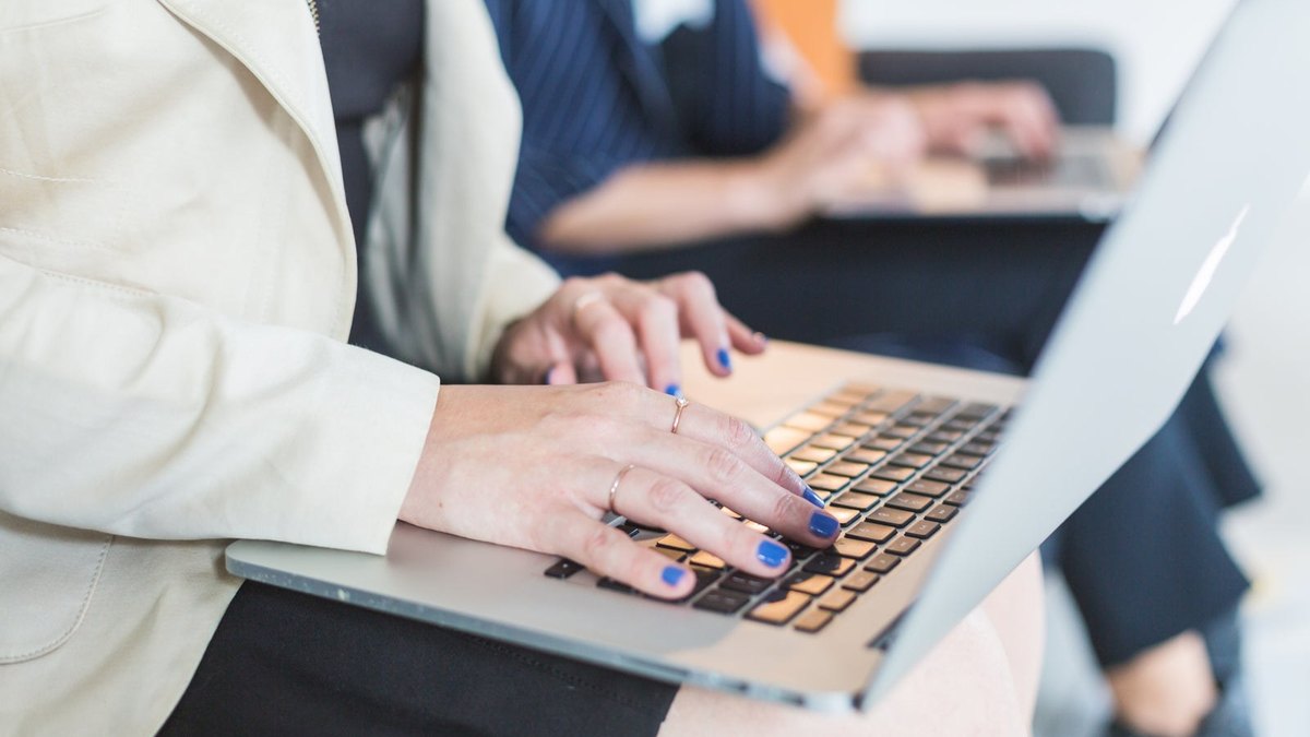 Close-up of hands typing on a laptop with another person working in the background.