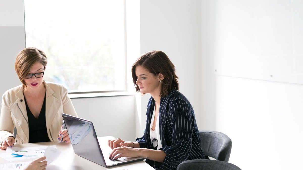 Two people reviewing a laptop together at a table beside a bright window.