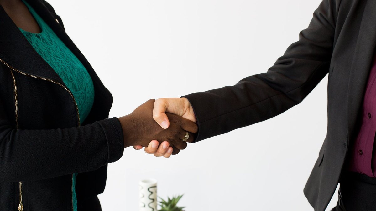 Two people in dark jackets shaking hands against a pale office background.
